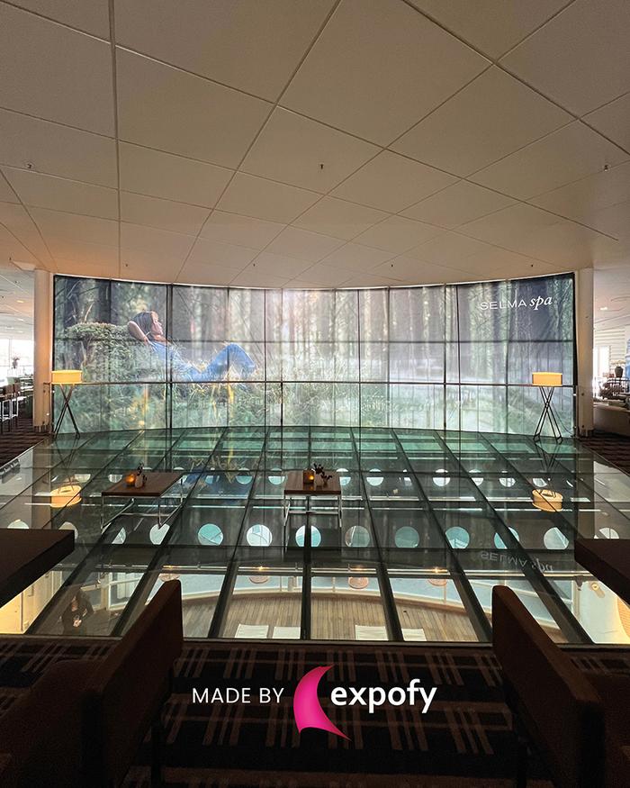 Stor kurvad bildvägg med skogslandskap och Selma Spa-logotyp i hotelllobby med glasgolv och atrium
