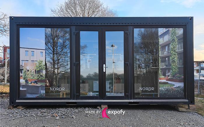Mörk containermodul med stora fönstergrafik på glasdörrar, bildtryck av bostadsmiljö och NORDR-logotyp, placerad utomhus.