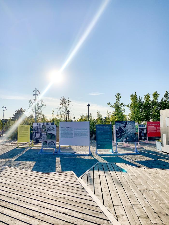 Utomhusutställning med fristående informationsskyltar med historiska bilder och texter på träbotten i solsken.