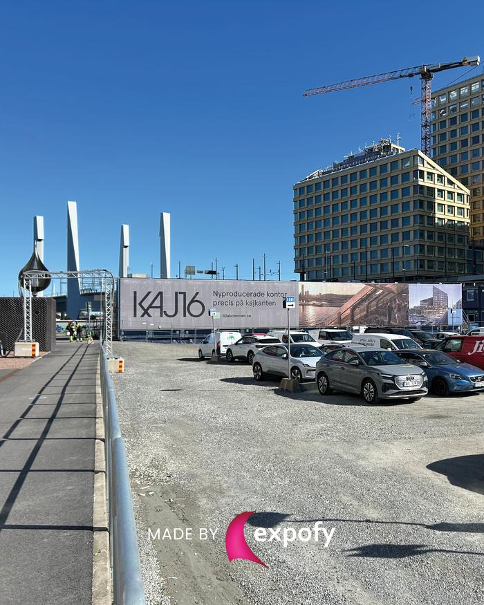Stor tryckt byggplanksbanner med KAJ16-logotyp framför en modern stadsmiljö med kran och höghus.