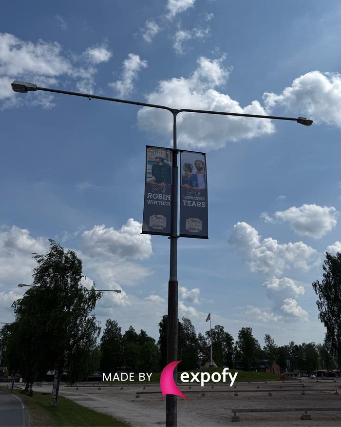 Två hängande gatubanners med porträttfotografier monterade på en gatulykta mot blå himmel med moln.