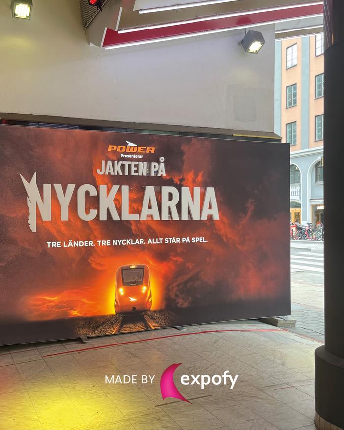 Stor tryckt backdrop med motiv av tåg mot eldslågor och orange himmel. Text: Jakten på Nycklarna. Power-logotyp.