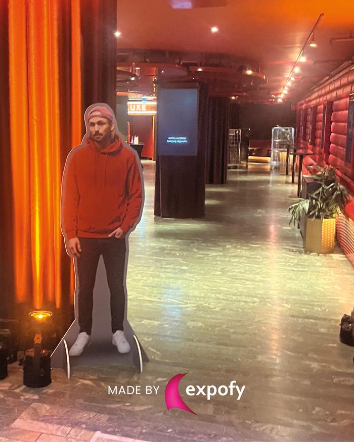 Livsstor utskuren figur av man i röd hoodie placerad i en mörkare inomhusmiljö med orange belysning.
