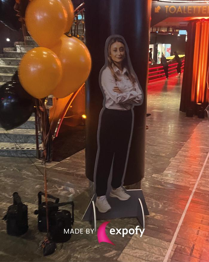 Lifesize-utklipp av kvinna med korslagda armar, placerat vid en pelare med orange och svarta ballonger i bakgrunden.