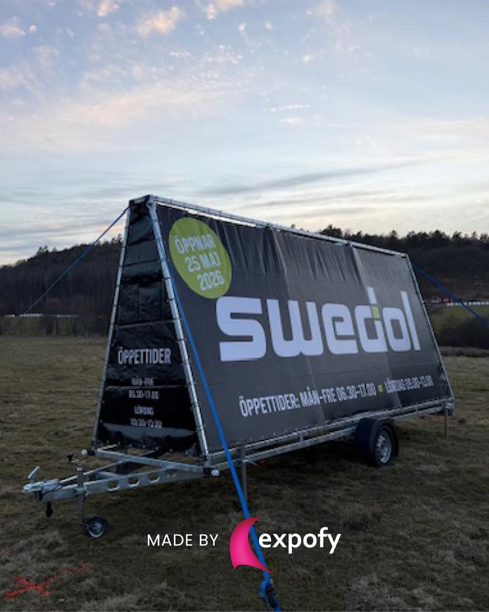 Tryckt reklamvepa på trailer med Swedol-logotyp, öppettider och öppningsdatum, parkerad på ett fält.